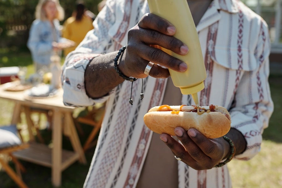 hands of young black man pouring sauce over hot dog in outdoor party